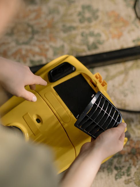 A person holding a yellow portable carpet cleaning machine with a black vent cover, positioned over a patterned carpet in a room with neutral lighting. The machine's handle and control panel are visib