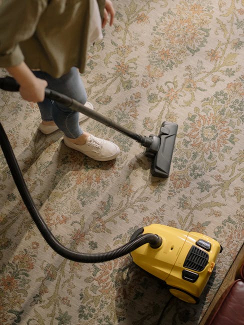 A person holding a yellow portable carpet cleaning machine with a black vent cover, positioned over a patterned carpet in a room with neutral lighting. The machine's handle and control panel are visib
