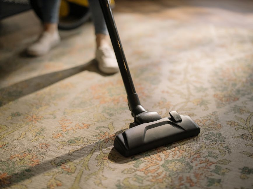 A young boy vacuuming the carpet in a bright, well-lit living room with a smiling man sitting on a cream-colored sofa, holding a book. The room features a wooden floor with a yellow and white patterne